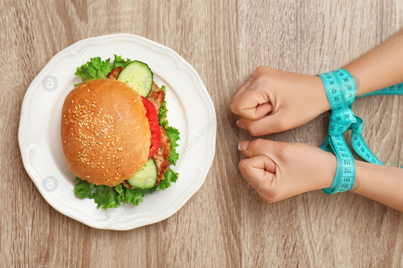 Photo of Woman holding tied hands with measuring tape near tasty sandwich on wooden table, top view. Healthy diet