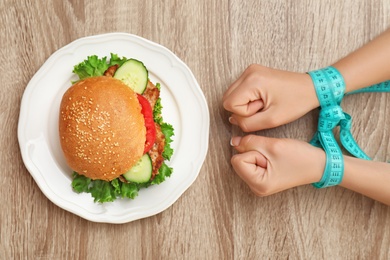 Woman holding tied hands with measuring tape near tasty sandwich on wooden table, top view. Healthy diet