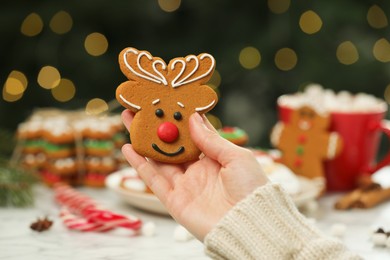 Woman with decorated cookie at table against blurred Christmas lights, closeup