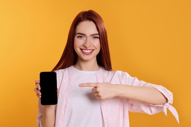 Photo of Happy woman with red dyed hair pointing at phone on orange background