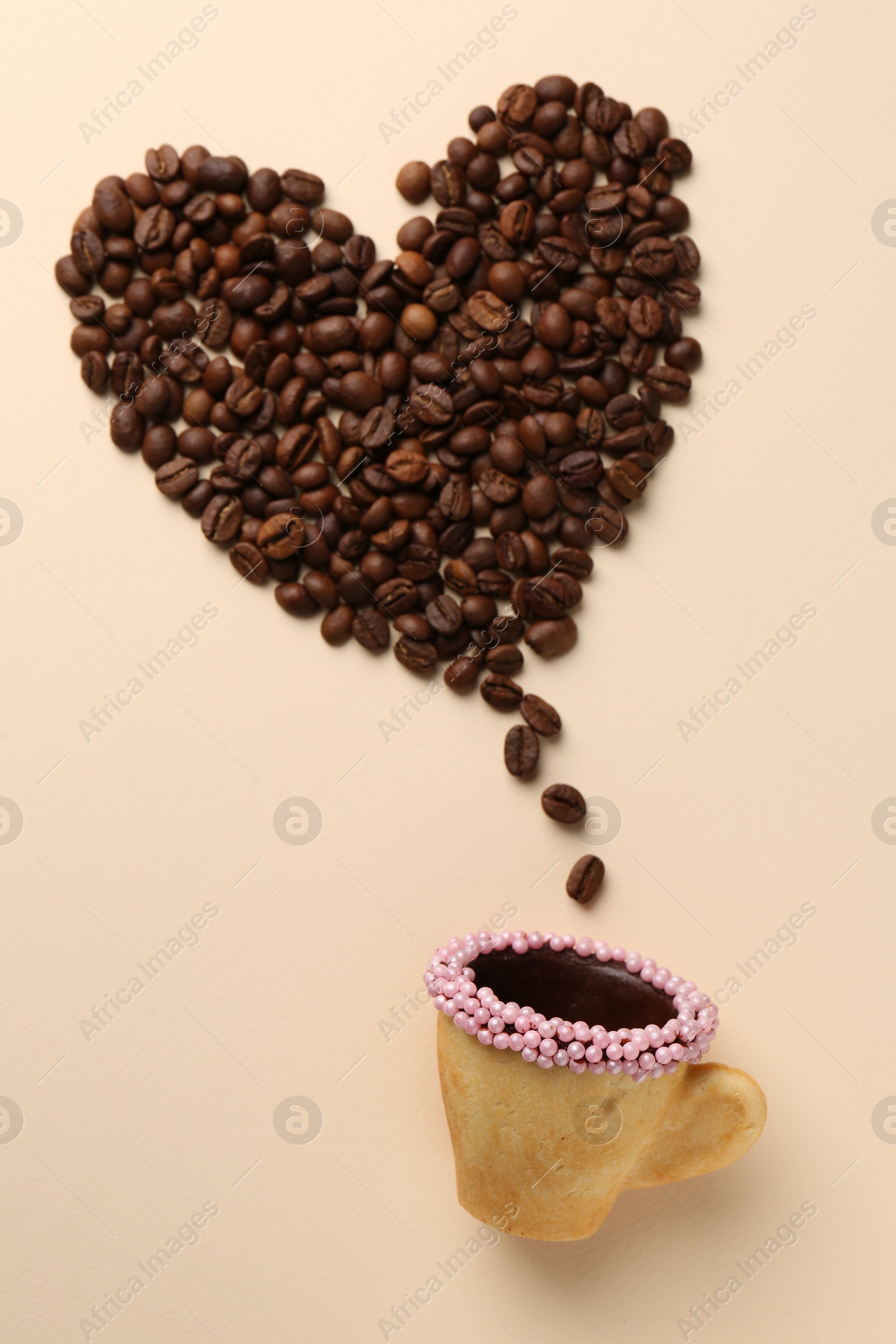 Photo of Coffee beans spilled from edible biscuit cup in shape of heart on beige background, top view