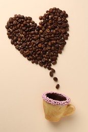 Photo of Coffee beans spilled from edible biscuit cup in shape of heart on beige background, top view