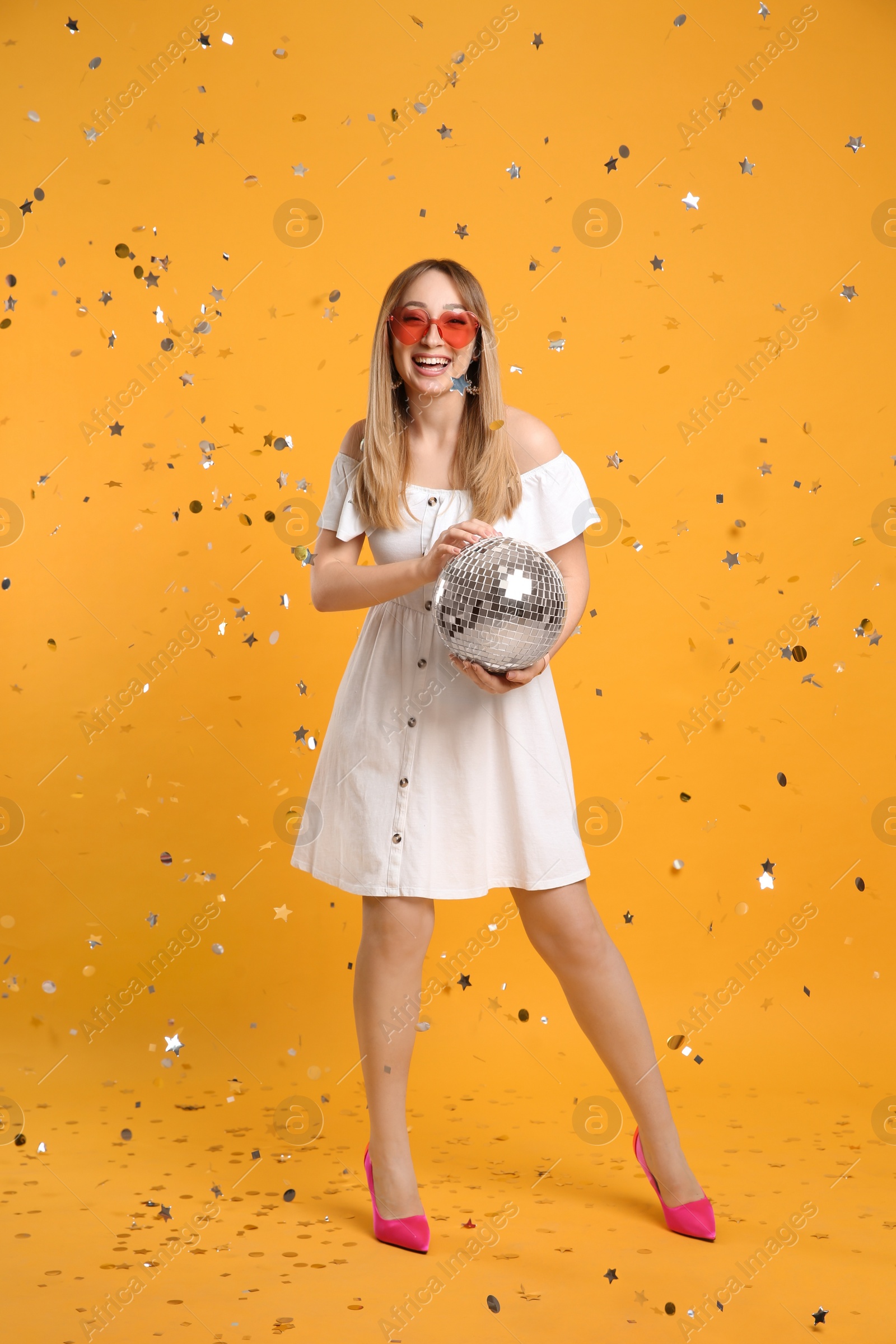 Photo of Happy young woman with disco ball and confetti on yellow background