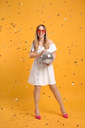 Happy young woman with disco ball and confetti on yellow background