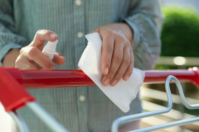 Photo of Woman cleaning handle of shopping cart with wet wipe and antibacterial spray on blurred background, closeup