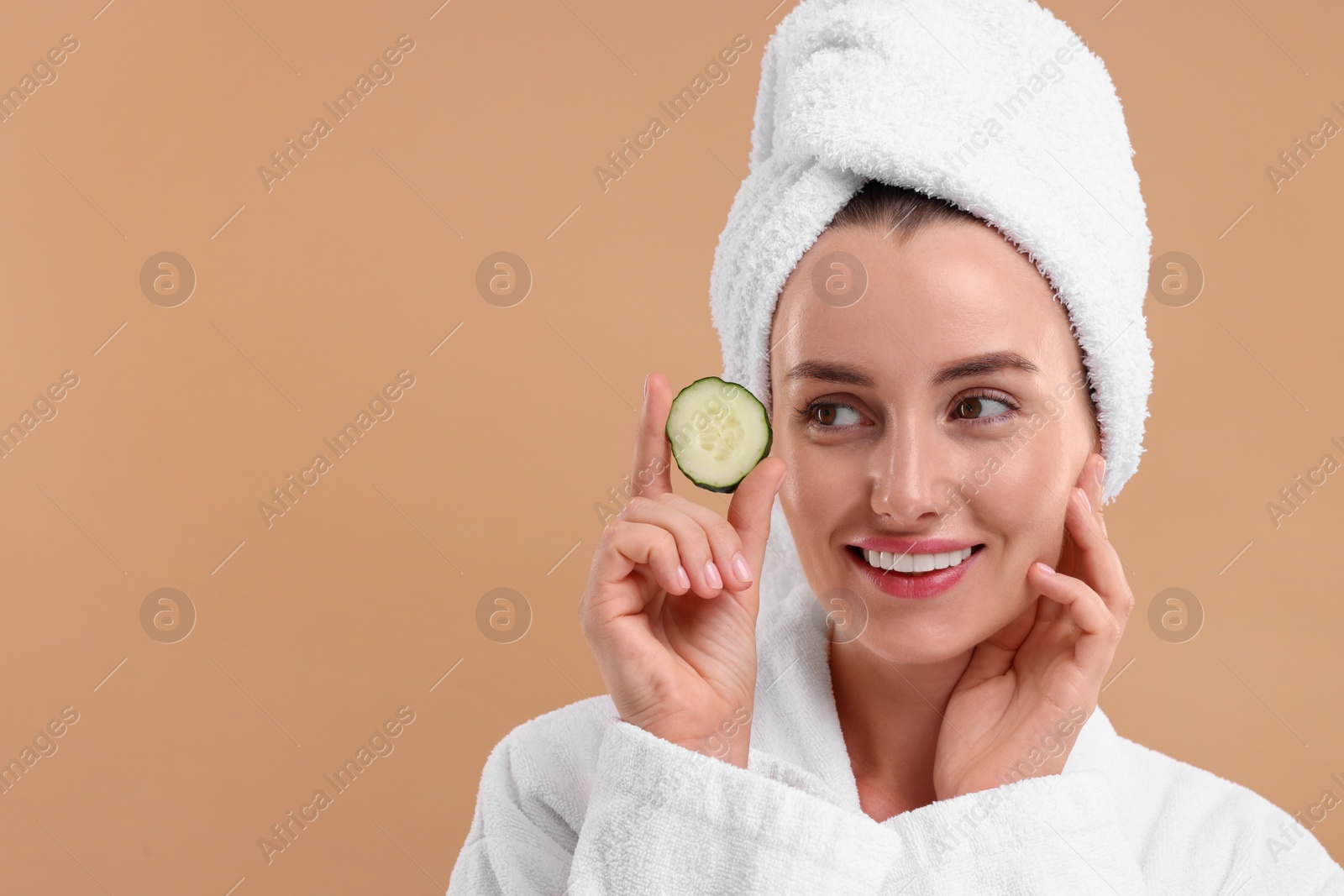 Photo of Beautiful woman in bathrobe with piece of cucumber on beige background, space for text