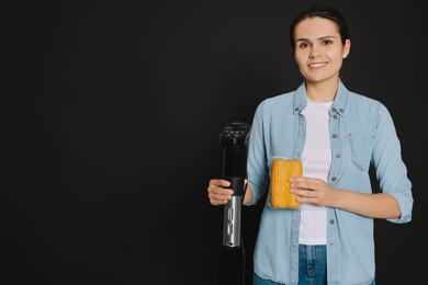 Photo of Beautiful young woman holding sous vide cooker and corn in vacuum pack on black background. Space for text