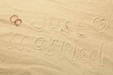 Honeymoon concept. Two golden rings and phrase Just married written on sand, top view