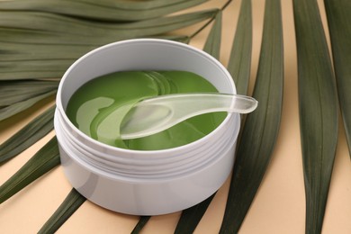 Photo of Jar of under eye patches with spoon and palm leaves on beige background, closeup. Cosmetic product