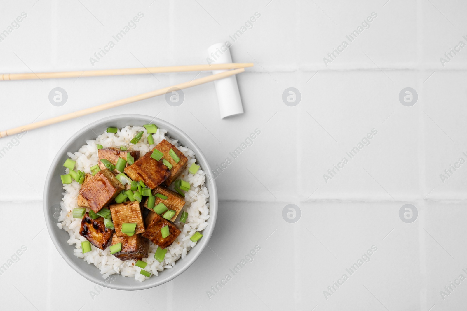 Photo of Bowl of rice with fried tofu and greens on white tiled table, flat lay. Space for text