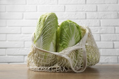 Photo of Fresh Chinese cabbages in string bag on wooden table near white brick wall