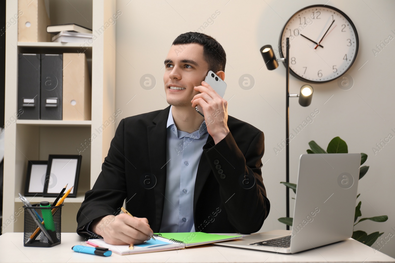 Photo of Man taking notes while talking on smartphone at table in office