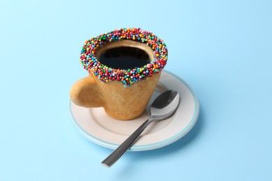 Photo of Delicious edible biscuit cup of coffee decorated with sprinkles and spoon on light blue background
