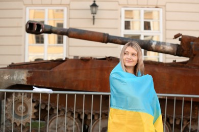 Photo of Young woman wrapped in Ukrainian flag near broken tank on street