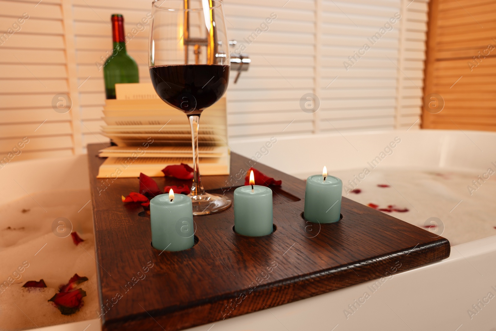 Photo of Wooden board with glass of wine, book, burning candles and rose petals on bath tub