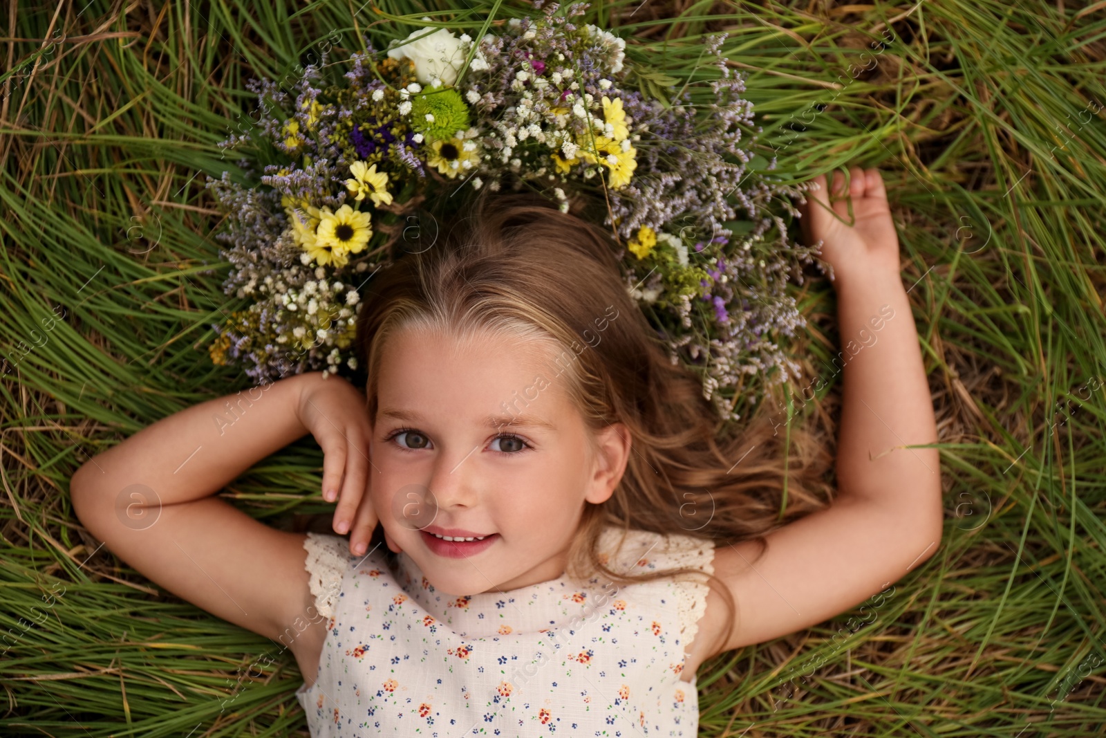 Photo of Cute little girl wearing wreath made of beautiful flowers on green grass, top view