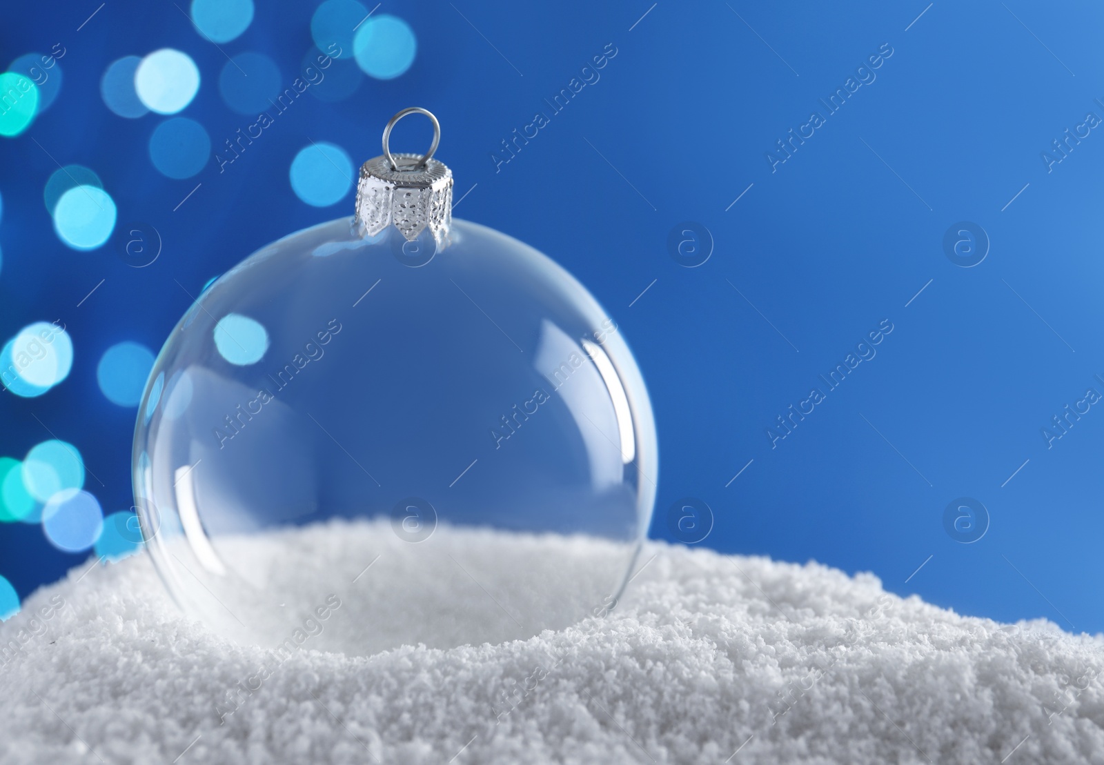 Photo of Transparent Christmas ball on snow against blurred fairy lights, space for text