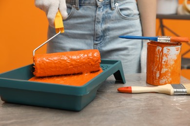 Photo of Woman taking orange paint with roller from tray at table, closeup