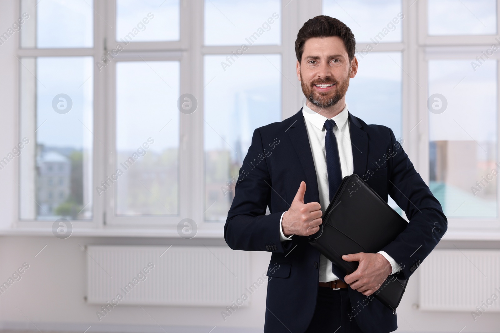 Photo of Happy real estate agent with leather portfolio showing thumb up in new apartment. Space for text