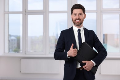 Happy real estate agent with leather portfolio showing thumb up in new apartment. Space for text