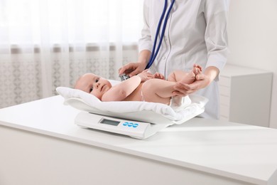 Pediatrician weighting and examining cute little baby with stethoscope in clinic, closeup