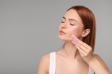 Young woman massaging her face with rose quartz gua sha tool on grey background, space for text