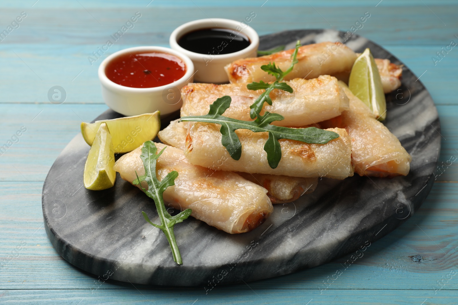 Photo of Tasty fried spring rolls, arugula, lime and sauces on light blue wooden table, closeup