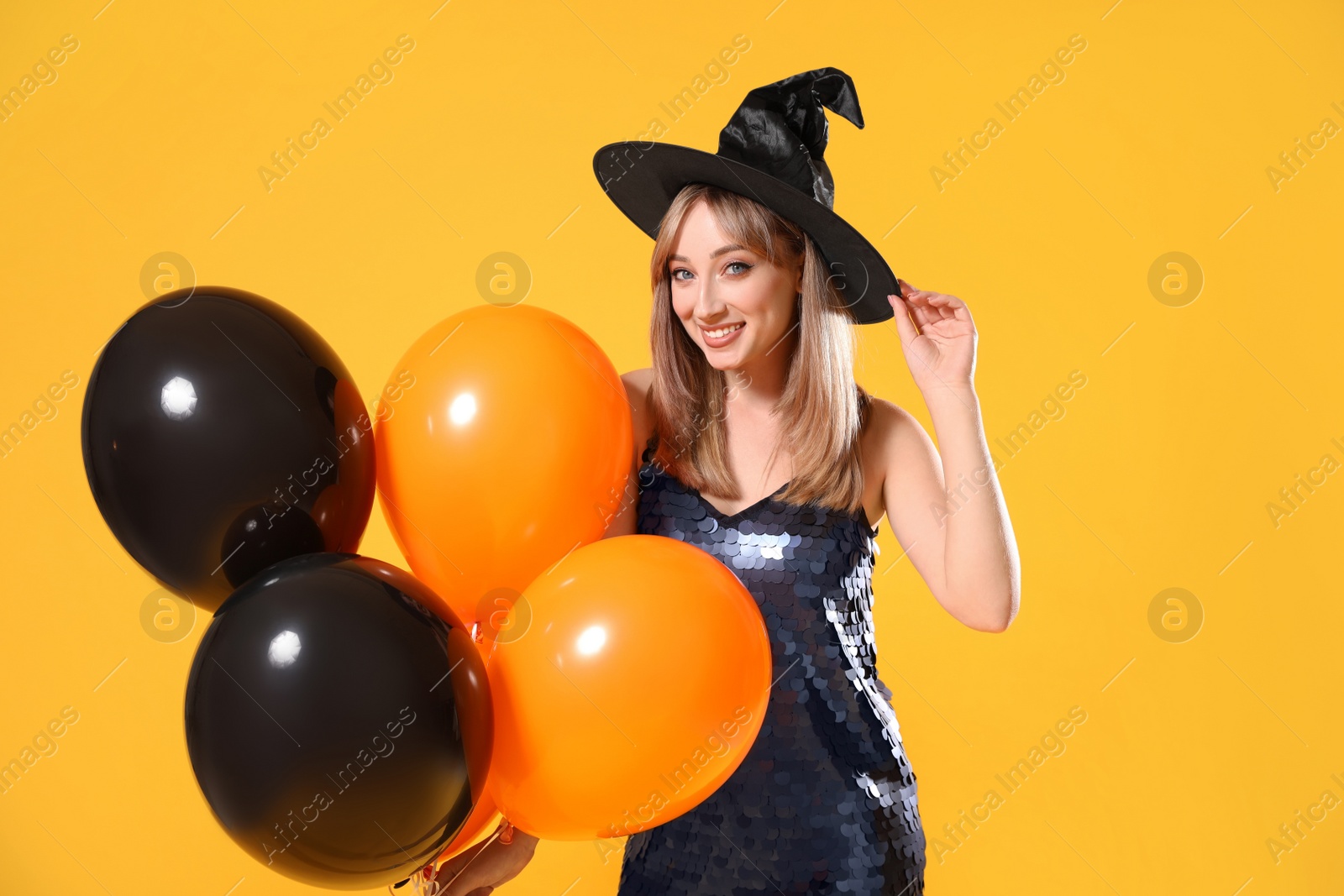 Photo of Beautiful woman in witch costume with balloons on yellow background. Halloween party