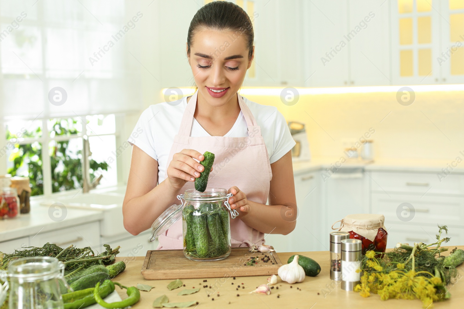 Photo of Woman putting cucumber into pickling jar at table in kitchen