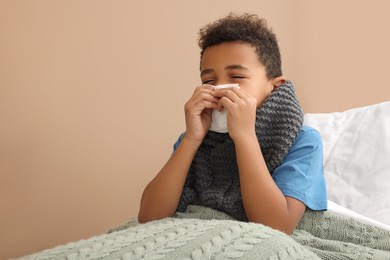 Photo of African American boy with scarf and tissue blowing nose in bed indoors, space for text. Cold symptoms