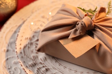 Photo of Furoshiki technique. Gift packed in beige fabric, with blank card on plate, closeup