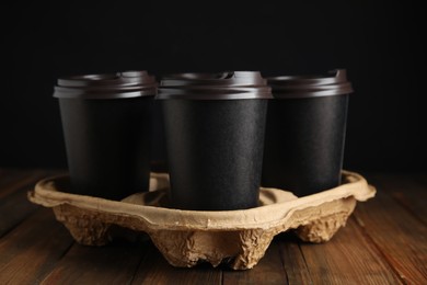 Photo of Takeaway paper coffee cups in cardboard holder on wooden table against black background