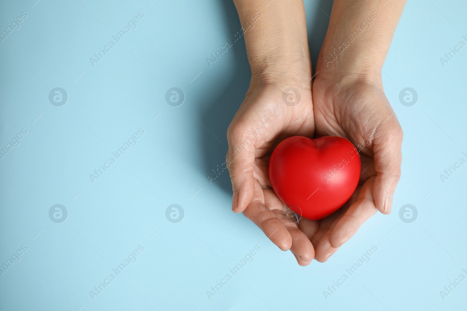 Photo of Elderly woman holding red heart in hands on light blue background, top view. Space for text