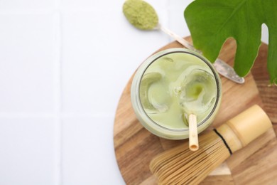 Photo of Glass of tasty iced matcha latte, leaf, bamboo whisk and powder on white tiled table, flat lay. Space for text