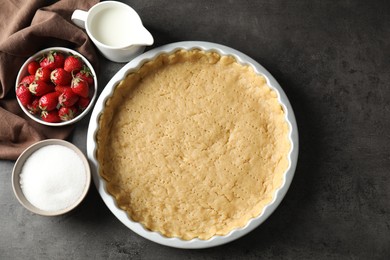 Photo of Making shortcrust pastry. Raw dough in baking dish, milk, sugar and strawberries on grey table, top view
