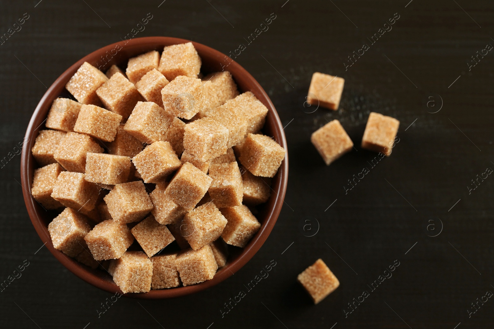 Photo of Brown sugar cubes in bowl on black wooden table, top view