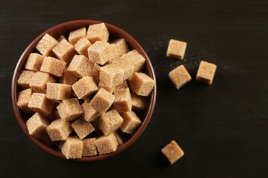 Photo of Brown sugar cubes in bowl on black wooden table, top view