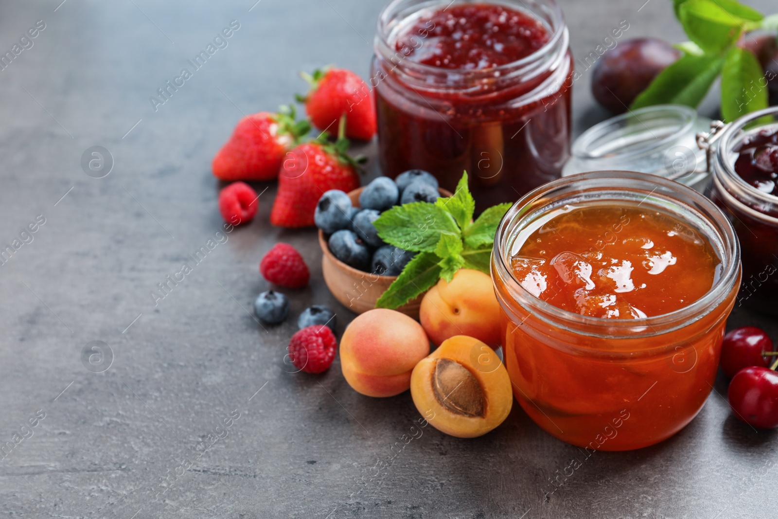 Photo of Jars with different jams and fresh fruits on grey table. Space for text