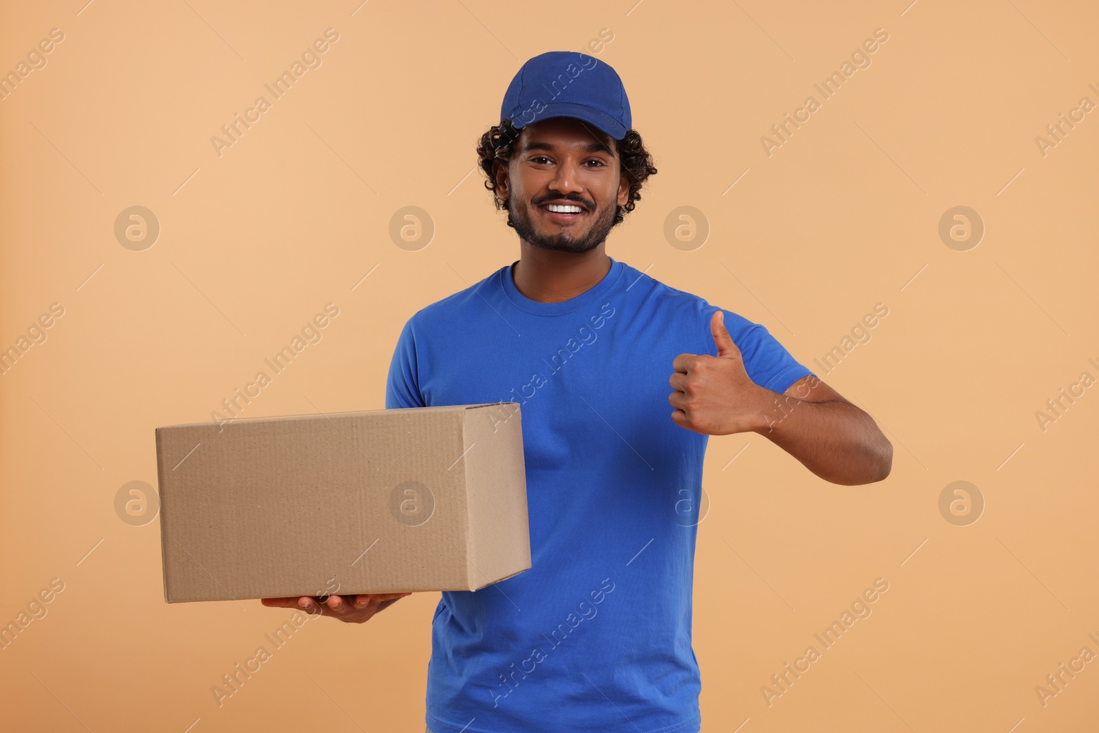 Photo of Happy young courier with parcel showing thumb up on light brown background