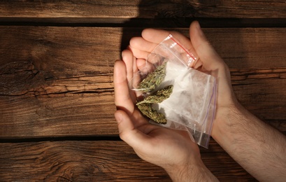 Photo of Man holding plastic bags with cocaine and hemp buds on wooden background, top view. Space for text