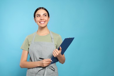 Young woman in grey apron with clipboard on light blue background, space for text