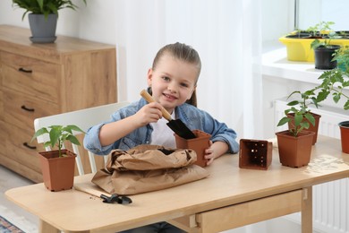 Cute little girl planting seedling into pot at wooden table in room