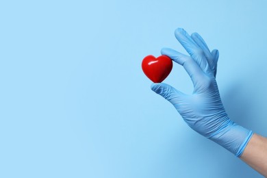 Photo of Doctor wearing medical glove holding decorative heart on light blue background, closeup. Space for text