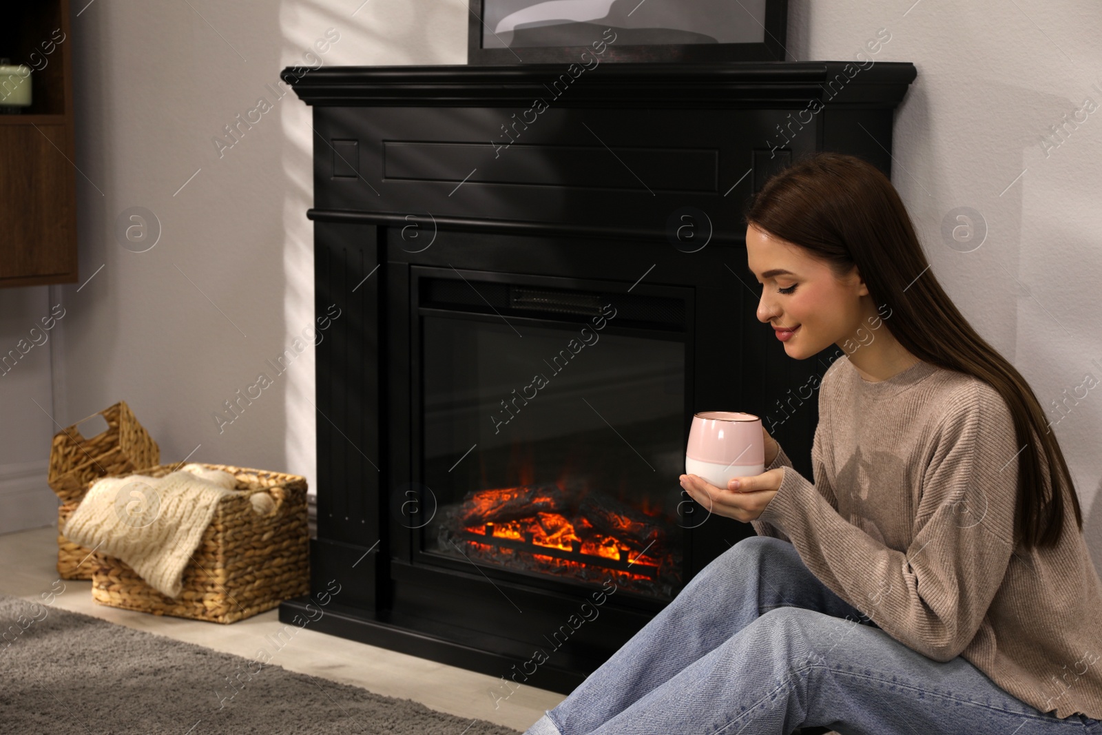 Photo of Beautiful young woman with cup of hot drink sitting on floor near fireplace at home. Space for text