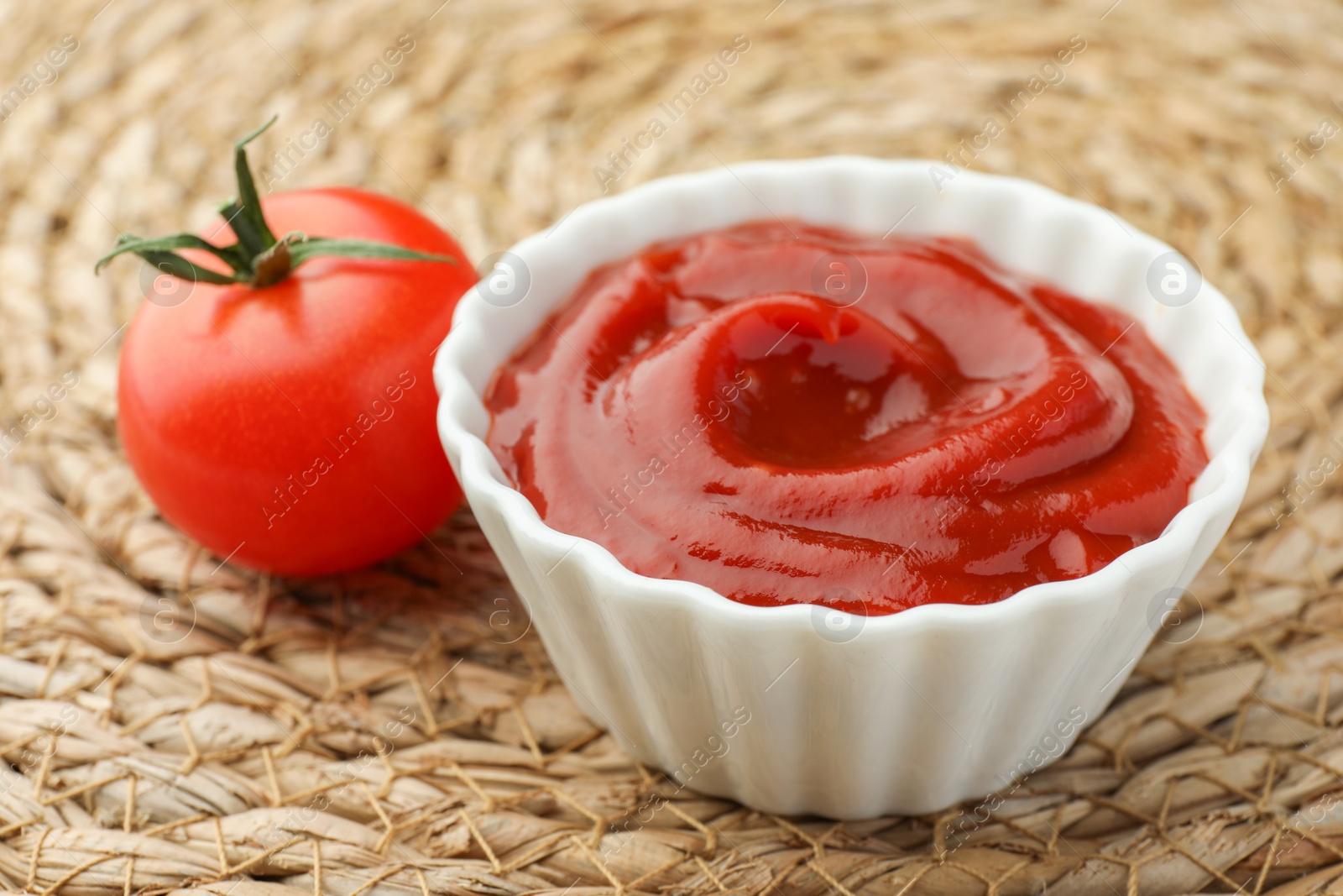 Photo of Bowl of tasty ketchup and tomato on wicker mat, closeup