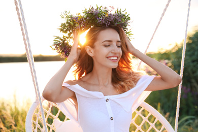 Young woman wearing wreath made of beautiful flowers on swing chair outdoors at sunset