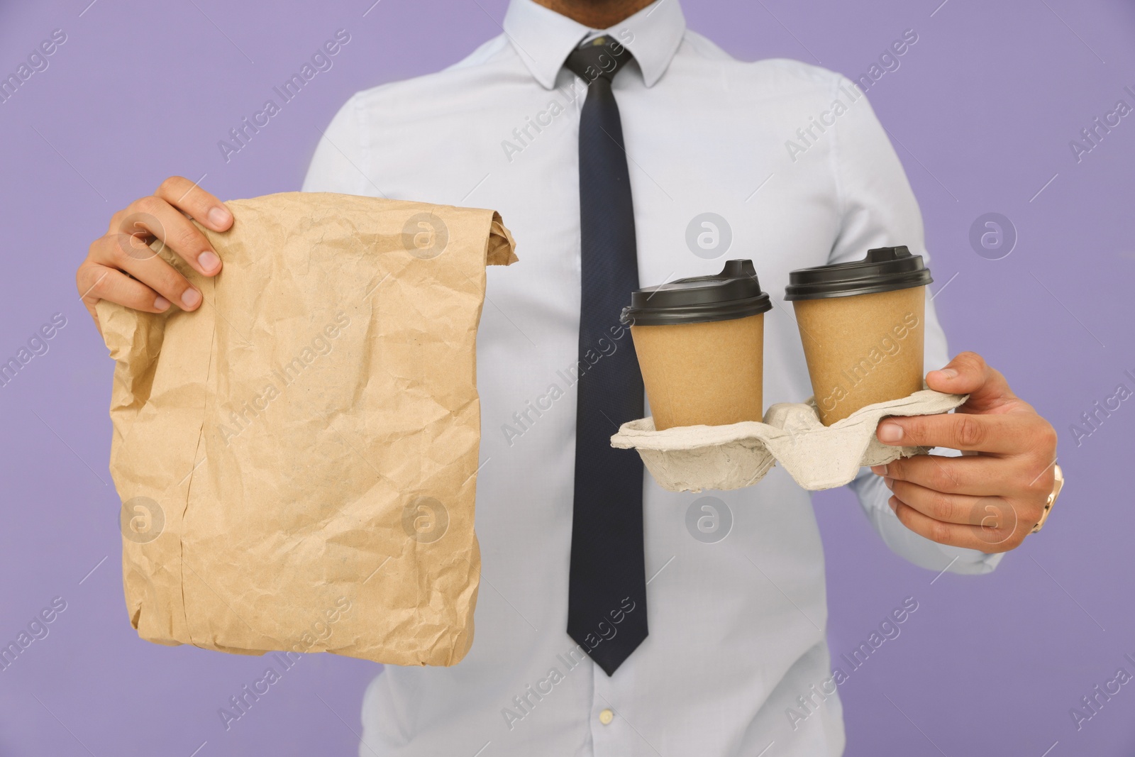 Photo of Young intern holding takeaway cups with hot drink and paper bag on lilac background, closeup
