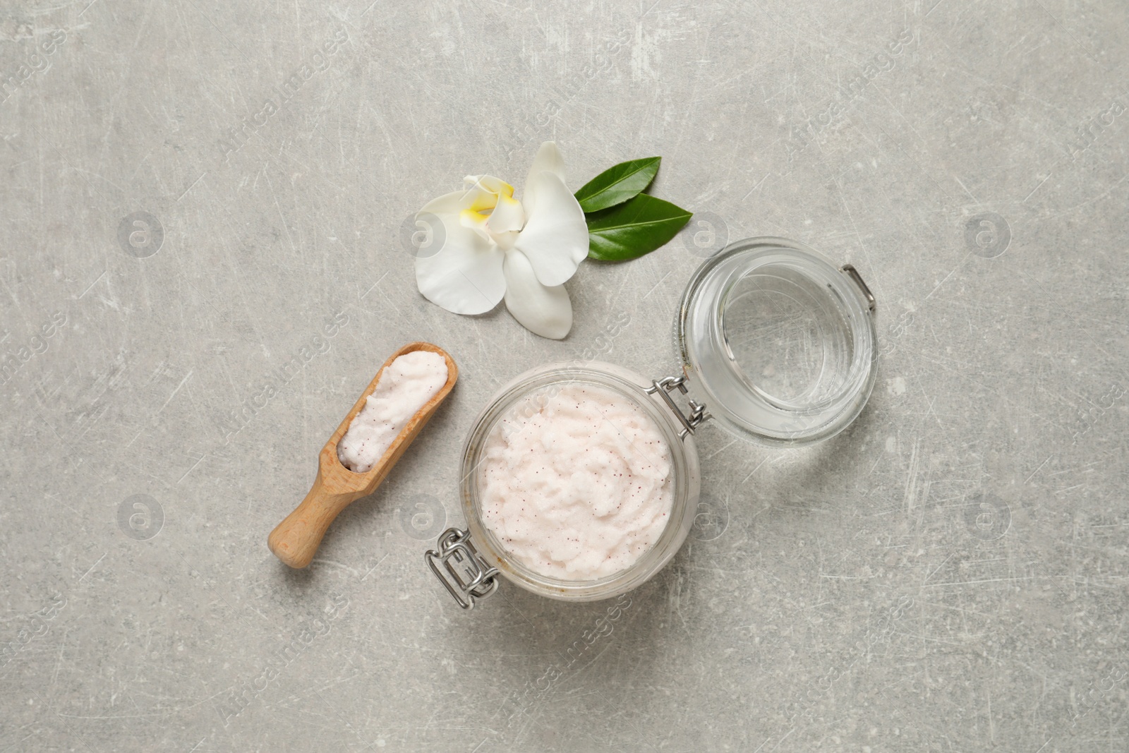 Photo of Body scrub in glass jar and scoop near orchid flower on light grey table, flat lay