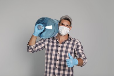 Photo of Courier in medical mask with bottle for water cooler showing thumb up on light grey background. Delivery during coronavirus quarantine