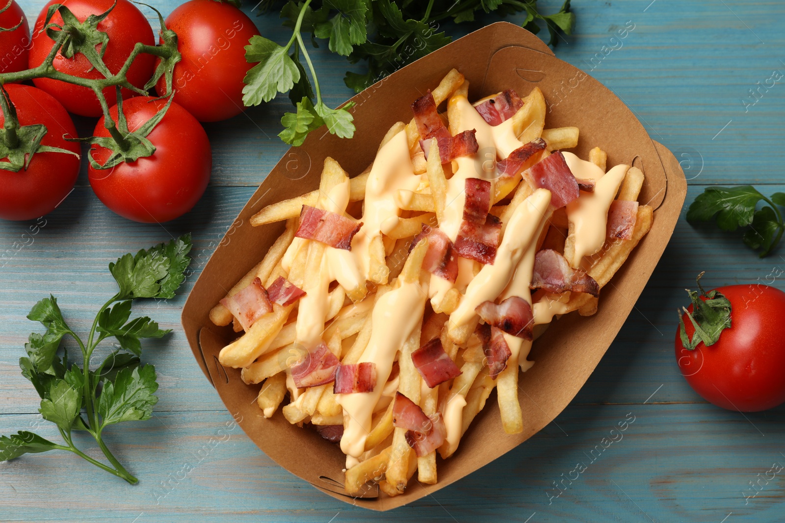 Photo of Tasty potato fries, cheese sauce, bacon and products in paper container on light blue wooden table, top view